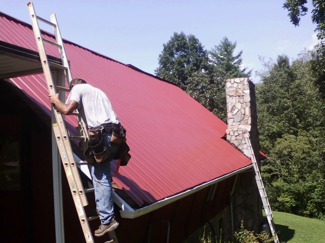 Roofer on a ladder beside a red metal roof and stone chimney — Soffit & Fascia Repair by Roofing Brothers in Walthourville